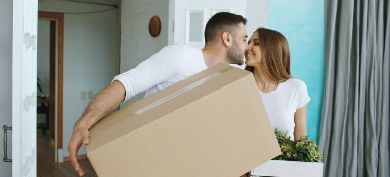 Happy couple moving into new home, carrying a box and smiling.